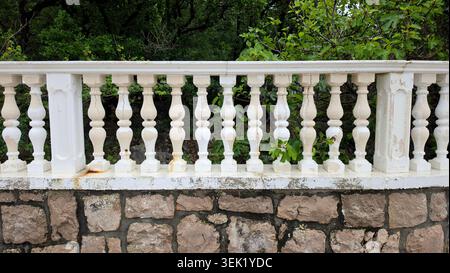 A peeling white classic balustrade fence stands on a rustic stone block wall with green common fig tree leaves behind it Stock Photo