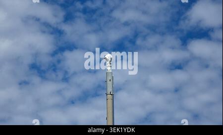 A low-angle shot captures a white motorized ptz surveillance camera fixed atop a high metal and concrete pole under a bright blue sky Stock Photo