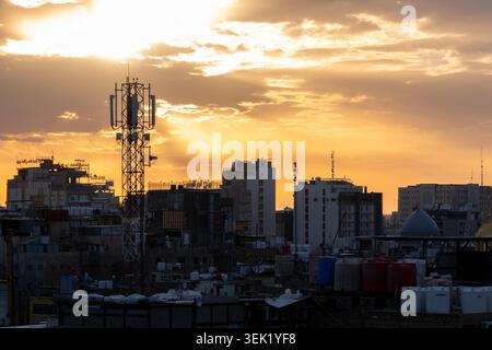 Karbala, Iraq - December 25, 2025: Urban skyline at sunset with a telecommunications tower rising above city buildings under a dramatic golden sky. Stock Photo