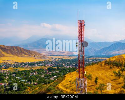 Tall red and white lattice communication tower stands on a golden hillside, overlooking a town in a valley. The scene features green trees and majestic mountains under a clear blue sky. Stock Photo