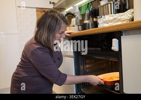 Woman prepares a homemade meal, carefully placing a dish with food into a hot oven, demonstrating domestic life and cooking activities in a cozy Stock Photo