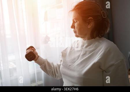 Senior woman exercising at home, lifting a dumbbell by a sunlit window, representing healthy aging, strength training, and an active lifestyle indoors Stock Photo