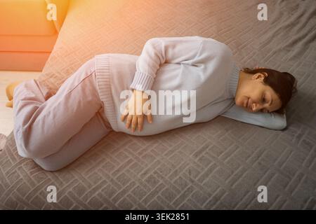 Pregnant woman resting on a bed in a softly lit bedroom, lying on her side, relaxed expression, cozy sweater and pants, warm morning light across the Stock Photo