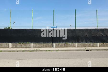 Road directional arrow sign on a concrete sidewalk. On behind a fence consisting of black tarpaulin with high wire mesh. Road in front. Background Stock Photo