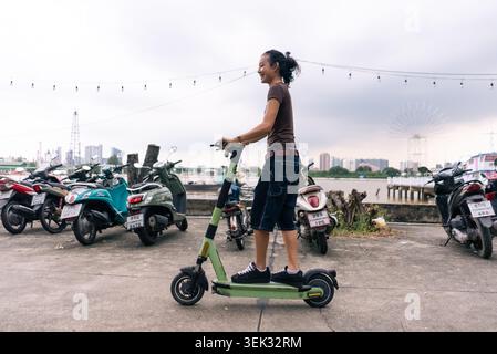 Young man riding electric scooter on pavement, smiling, next to river in urban setting Stock Photo