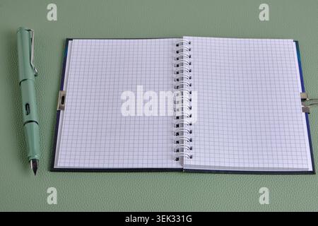 Green fountain pen next to an open blank spiral checkered notebook on matching textured surface Stock Photo