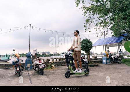 Young man riding electric scooter along tropical city waterfront Stock Photo