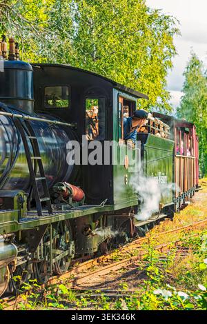 Jokioinen, Finland - 29.08.2020: Historic steam train with engineer in cab emitting white smoke on tracks amidst bright summer foliage Stock Photo