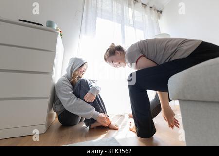 Young woman in gray hoodie sits on floor while another woman kneels beside her, both in a bright room with natural light and modern furniture Stock Photo