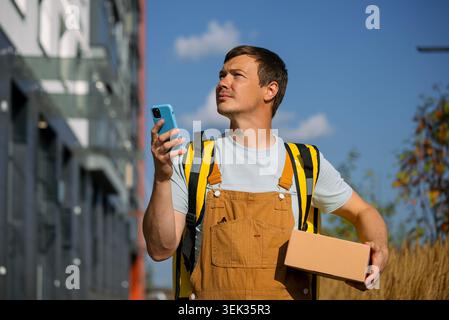 Delivery courier in yellow backpack and workwear holds a cardboard box and smartphone, looking to the side on a city street, symbolizing urban logistics and real-time delivery Stock Photo