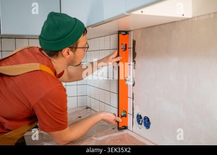 Worker carefully installing ceramic tiles on a kitchen backslash, ensuring perfect horizontal and vertical alignment using a bubble level for precise home improvement Stock Photo