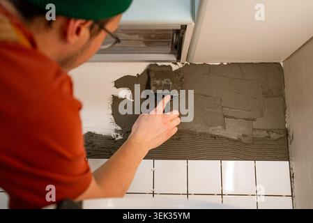 Construction worker applying tile adhesive to a kitchen wall with a trowel, preparing for the installation of new ceramic tiles on the backsplash during a home renovation project Stock Photo