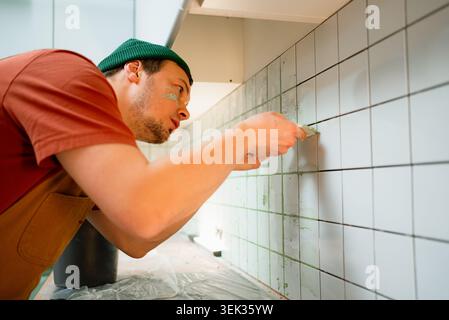 Concentrated young man works on a home improvement project, carefully applying grout to a white tiled kitchen backsplash. Side view shot with shallow depth of field. Concept of DIY and renovation. Stock Photo
