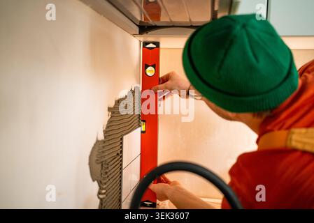 Tiler applying tile adhesive and carefully checking the alignment of new ceramic tiles with a spirit level on a kitchen backsplash wall, actively working on home renovation Stock Photo