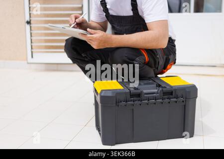 Professional technician using a digital tablet and stylus to document repair work against a modern backdrop, sitting on his toolbox while performing a Stock Photo