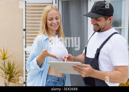 Female customer smiling and interacting with a delivery man in uniform, signing for received goods on a digital tablet using a stylus at her residenti Stock Photo