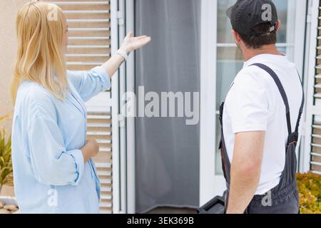 Woman standing outside her house pointing at a window shutter while explaining repair needs to a technician during a home maintenance consultation on Stock Photo