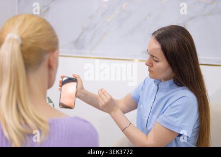 Female cosmetologist expert discussing client skin condition with mobile phone ai dermatoscopy analysis, advising on modern skincare treatments in bea Stock Photo