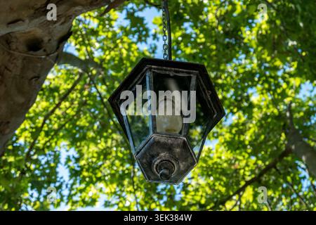 A vintage black metal hexagonal hanging lantern suspended from a chain among lush green tree canopy against a blue sky, Saint Quinten la Poterie Stock Photo