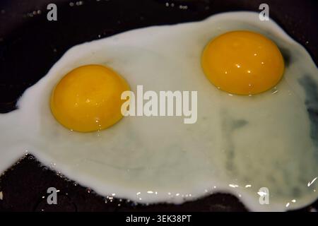 Close up shot of two fresh fried eggs cooking in a black non-stick frying pan. The vibrant, glossy yellow egg yolks are liquid and perfectly intact, s Stock Photo