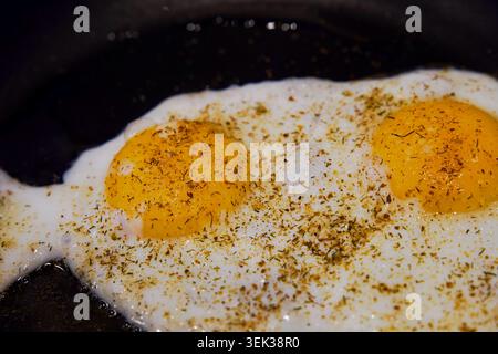High angle view of two sunny-side-up fried eggs seasoned with dried green herbs and spices cooking in a black frying pan. The glossy yellow yolks are Stock Photo