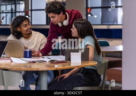 Diverse students collaborating in study lounge around round table with laptop and textbook Stock Photo