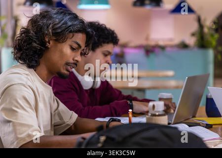 Indian male friends writing notes and working on laptop at cafe table with notebooks, pen, coffee Stock Photo