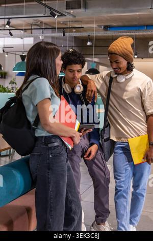 Diverse students in study lounge, viewing tablet, holding red and yellow folders with headphones Stock Photo