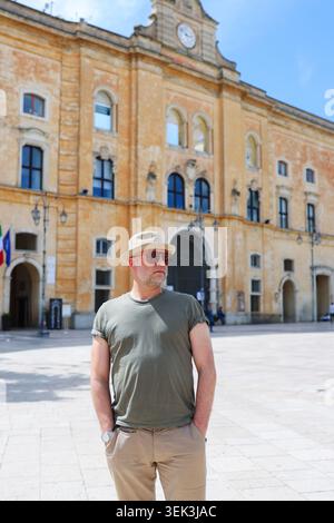 Stylish middle-aged man in sunglasses and hat standing in a historic Italian square, enjoying sunny travel and cultural tourism in Europe Stock Photo