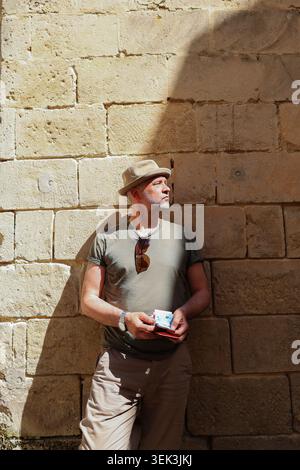 Modern tourist with hat and smartphone relaxing by ancient wall in Mediterranean city with dramatic light and shadows. Stock Photo