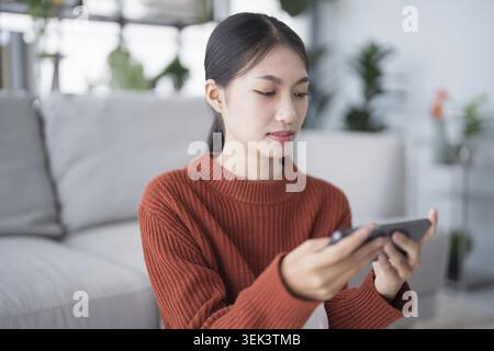 Asian woman controls smart home. Mobile phone with smart home app in the living room. Touch screen AI technology. Remote control system for your home Stock Photo