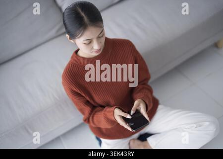 Asian woman controls smart home. Mobile phone with smart home app in the living room. Touch screen AI technology. Remote control system for your home Stock Photo