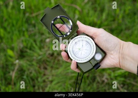 Hand holds an open military magnetic compass against a background of green grass. Equipment for hiking, studying geography, or tactical army training. Stock Photo