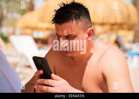 Caucasian man uses a smartphone at a beach resort in Sharm El Sheikh Egypt with wet hair and sunny outdoor setting. Remote work and mobile lifestyle. Stock Photo