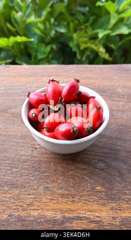 Fresh Rose Hips in White Bowl on Rustic Wooden Table with Natural Green Background Stock Photo
