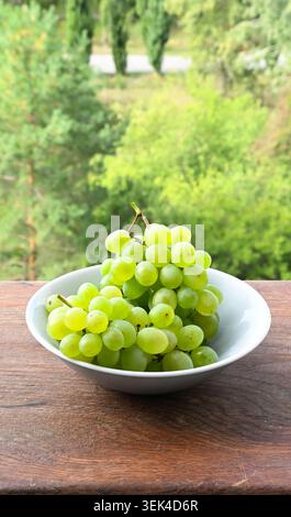Fresh green grapes in a white ceramic bowl on a rustic wooden table with a blurry nature background. Stock Photo