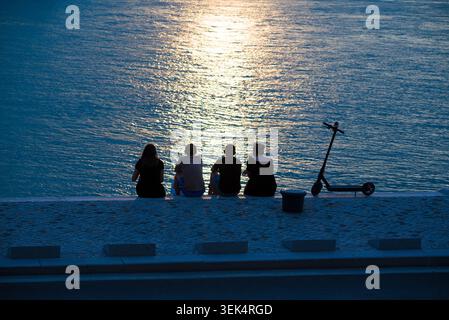 Four friends sitting together at dusk on a stone waterfront pier watching the sunset reflection over water, next to an electric kick scooter Stock Photo