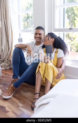 Diverse couple sitting on hardwood floor by windows, woman wearing yellow dress taking phone selfie Stock Photo