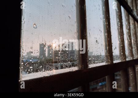 Raindrops on window screen with city skyline at sunset, urban view through mesh barrier Stock Photo