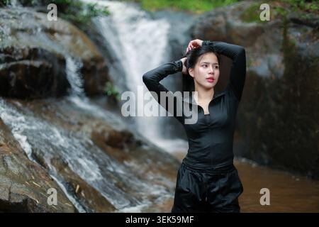 Young Asian woman wearing black athletic sportswear posing peacefully outdoors near natural rocky cascading waterfall showing confident serene express Stock Photo