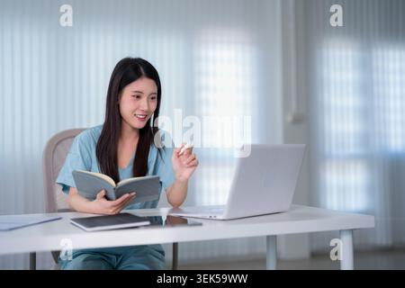 Asian woman reading notebook and using laptop computer at home office desk bright indoor workspace study learning Stock Photo