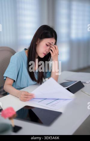stressed young Asian woman doctor reading medical report document feeling worried exhausted overwhelmed at desk Stock Photo