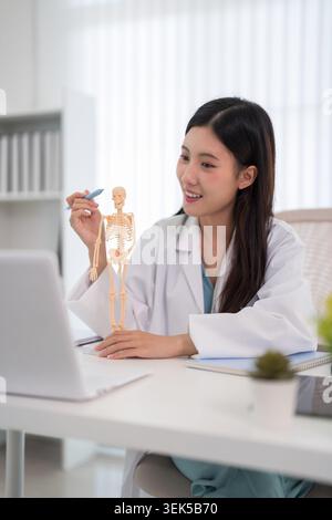 Young Asian woman doctor in white coat holding anatomical skeleton model during virtual medical education session Stock Photo