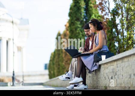 Young professionals collaborating outdoors on a laptop while seated on a stone wall in an urban park with clear, sunny weather and modern architecture Stock Photo