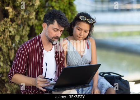 Man and woman collaborating outdoors on a laptop, engaged in focused teamwork by the water's edge Stock Photo