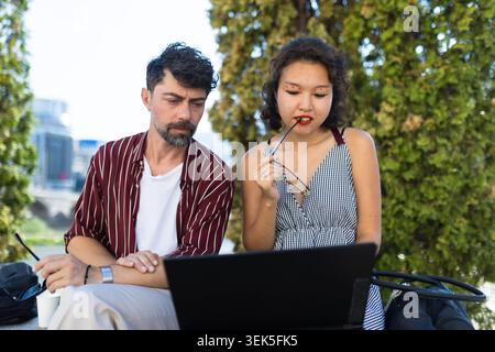 Young man and woman collaborating on a laptop outdoors with a thoughtful expression Stock Photo