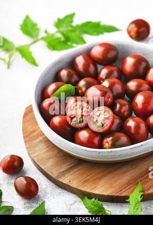 Fresh mini Kumato tomatoes (Solanum lycopersicum) in a ceramic bowl on a wooden board. Dark brown juicy cherry tomatoes, whole and halved. Stock Photo