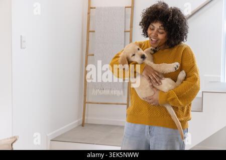 African American woman holding golden puppy on stair landing, wearing mustard sweater and jeans Stock Photo