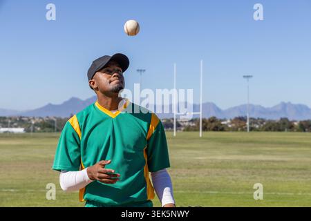African American man tossing baseball on grassy field wearing green jersey black cap white sleeve Stock Photo