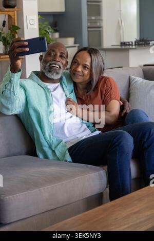Mature couple sitting on gray sofa at home taking selfie while man holds smartphone, wearing jeans Stock Photo